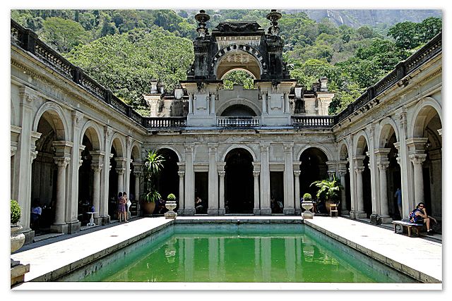 Visão interna do pátio da Escola de Artes Visuais do Parque Lage. A foto mostra uma grande construção em cores claras e um espelho d'água verde.